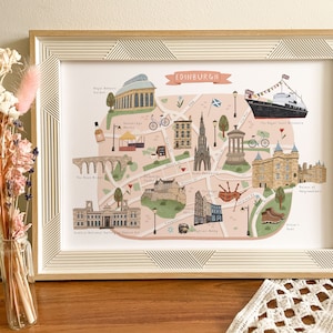 May include: A colorful illustrated map of Edinburgh, Scotland, with landmarks like Edinburgh Castle, the Royal Yacht Britannia, and the Sheep Held Inn. The map is framed in a light wood frame.