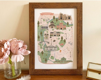 Puede incluir: Una ilustración enmarcada de un mapa de Cambridge, Inglaterra. El mapa es de color rosa claro con árboles verdes y edificios marrones. El texto "CAMBRIDGE" está en la parte superior del mapa.