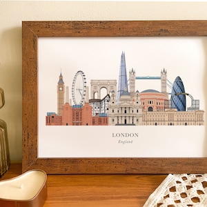Puede incluir: Una impresi&oacute;n enmarcada de una ilustraci&oacute;n del horizonte de Londres con monumentos emblem&aacute;ticos como el Big Ben, el London Eye, el Tower Bridge y el Shard. La impresi&oacute;n tiene un marco de madera marr&oacute;n y el texto "LONDON England" en la parte inferior.