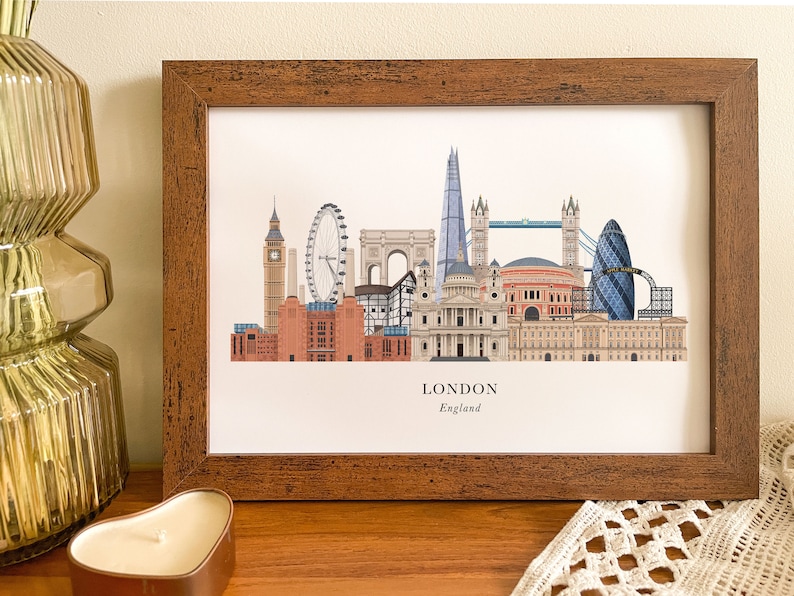 Puede incluir: Una impresi&oacute;n enmarcada de una ilustraci&oacute;n del horizonte de Londres con monumentos emblem&aacute;ticos como el Big Ben, el London Eye, el Tower Bridge y el Shard. La impresi&oacute;n tiene un marco de madera marr&oacute;n y el texto "LONDON England" en la parte inferior.