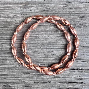 May include: A circular bracelet composed of polished copper-coloured metallic beads. The design features elongated, oval-shaped beads alternating with small, round spherical beads, creating a continuous, reflective, and uniform metallic loop.