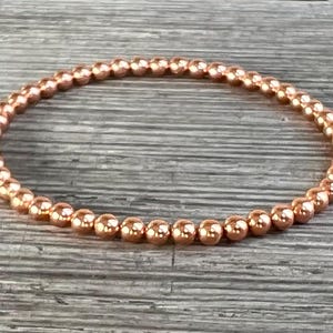 Op de afbeelding: Een koperkleurige kralenarmband. De armband is gemaakt van kleine, ronde kralen die aan elkaar zijn geregen om een flexibele band te vormen. De kralen hebben een glanzende, metallic afwerking. De armband wordt gepresenteerd op een houten oppervlak.
