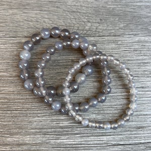 May include: Three bracelets made of grey, translucent beads. The bracelets are layered, with beads of varying sizes. The background is a wooden surface with a horizontal grain pattern.
