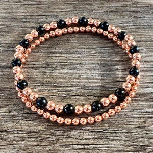 May include: Two beaded bracelets. One bracelet features small, rose gold-colored beads. The second bracelet has the same rose gold beads, interspersed with larger, black beads. The bracelets are displayed on a wooden surface.