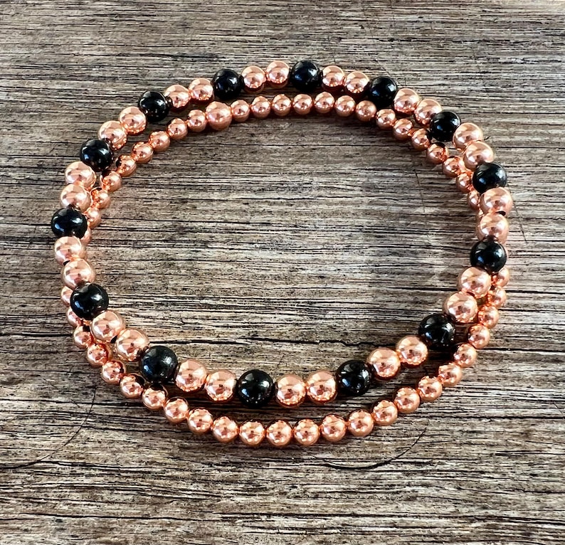 May include: Two beaded bracelets. One bracelet features small, rose gold-colored beads. The second bracelet has the same rose gold beads, interspersed with larger, black beads. The bracelets are displayed on a wooden surface.