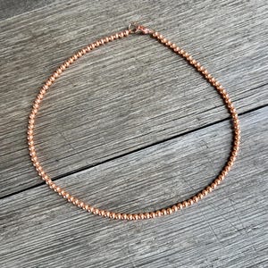 May include: A copper-colored beaded necklace with a clasp closure. The necklace is made of small, round beads and is approximately 18 inches in length.