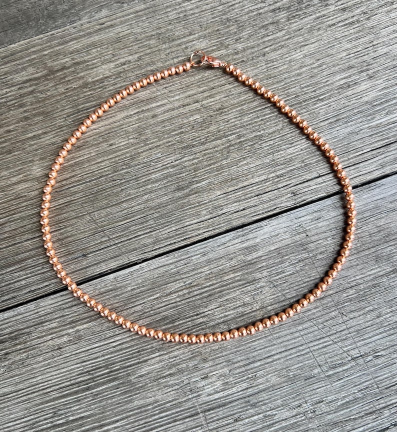 May include: A copper-colored beaded necklace with a clasp closure. The necklace is made of small, round beads and is approximately 18 inches in length.