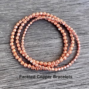 May include: Three strands of faceted copper bracelets are displayed on a wooden surface. The bracelets are a warm, reddish-brown color, with each bead reflecting light. The text "Faceted Copper Bracelets" is at the bottom.