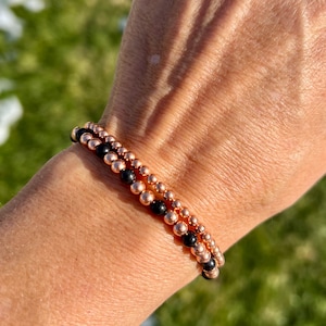 May include: Two beaded bracelets. One bracelet features small, black beads, while the other has small, rose gold-colored beads. The bracelets are stacked on top of each other. The bracelets are on a person's wrist.