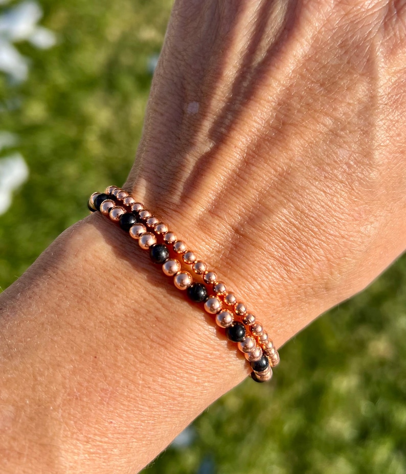 May include: Two beaded bracelets. One bracelet features small, black beads, while the other has small, rose gold-colored beads. The bracelets are stacked on top of each other. The bracelets are on a person's wrist.