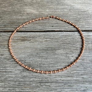 May include: A copper-coloured beaded necklace. The necklace features small, round beads strung together, creating a choker design. The beads have a metallic sheen, and the necklace is secured with a small clasp.