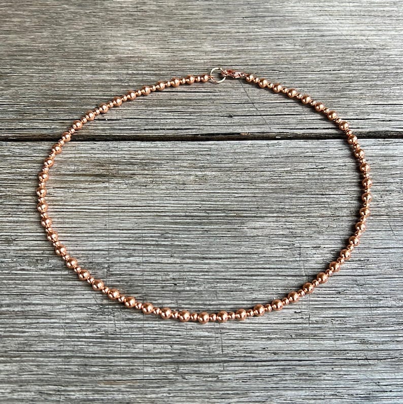May include: A copper-coloured beaded necklace. The necklace features small, round beads strung together, creating a choker design. The beads have a metallic sheen, and the necklace is secured with a small clasp.