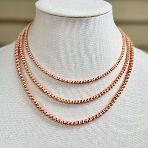 May include: Three rose gold necklaces displayed on a white mannequin. Each necklace is a different length, with a delicate beaded chain design. The necklaces are a warm, metallic color, and are suitable for layering.