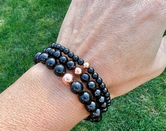 Puede incluir: Tres pulseras de cuentas negras con detalles en oro rosa. Las pulseras están apiladas en la muñeca de una persona.