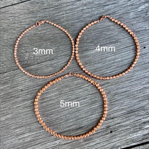 K&ouml;nnte beinhalten: Drei ros&eacute;goldene Perlenarmb&auml;nder mit einem Durchmesser von 3 mm, 4 mm und 5 mm werden auf einer Holzoberfl&auml;che gezeigt. Jedes Armband ist ein vollst&auml;ndiger Kreis mit einem Verschluss. Der Text "3mm", "4mm" und "5mm" ist aufgedruckt.