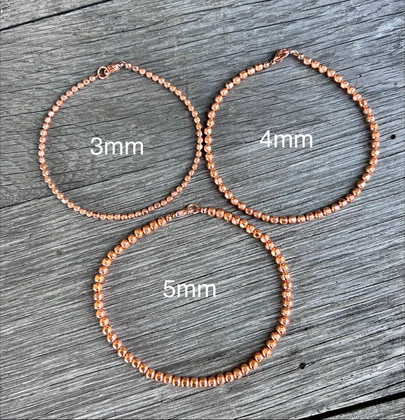 K&ouml;nnte beinhalten: Drei ros&eacute;goldene Perlenarmb&auml;nder mit einem Durchmesser von 3 mm, 4 mm und 5 mm werden auf einer Holzoberfl&auml;che gezeigt. Jedes Armband ist ein vollst&auml;ndiger Kreis mit einem Verschluss. Der Text "3mm", "4mm" und "5mm" ist aufgedruckt.