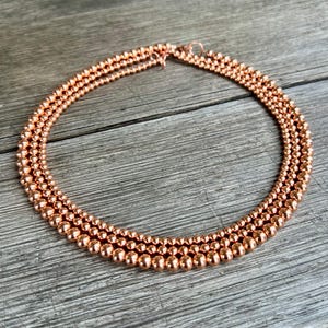 Puede incluir: Un collar de cobre con tres hileras de cuentas redondas. El collar está sobre una superficie de madera.