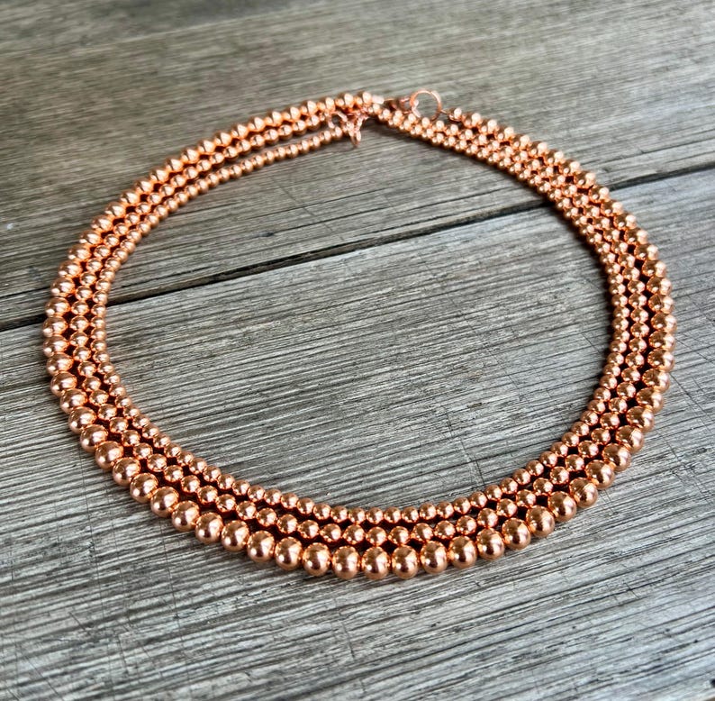 May include: A copper necklace with three strands of round beads. The necklace is on a wooden surface.