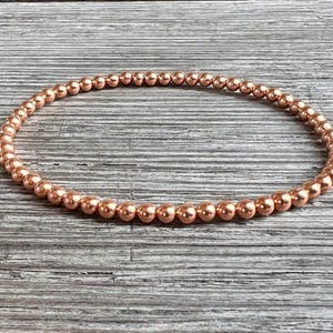 May include: A copper-coloured beaded bracelet. The bracelet is made of small, round beads strung together to form a flexible band. The beads have a metallic sheen and are evenly spaced. The bracelet is displayed on a wooden surface.
