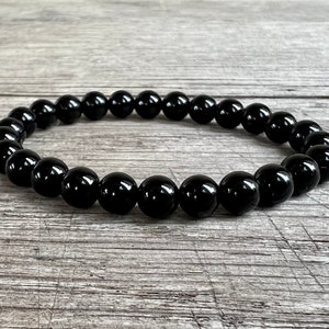 May include: A black beaded bracelet made of round, polished stones. The bracelet is lying on a light brown wooden surface.
