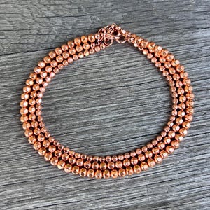 K&ouml;nnte beinhalten: Eine kupferfarbene Perlenkette mit dreireihigem Design. Die Halskette hat kleine, runde Perlen und einen passenden Verschluss. Der Schmuck wird auf einer Holzoberfl&auml;che pr&auml;sentiert.