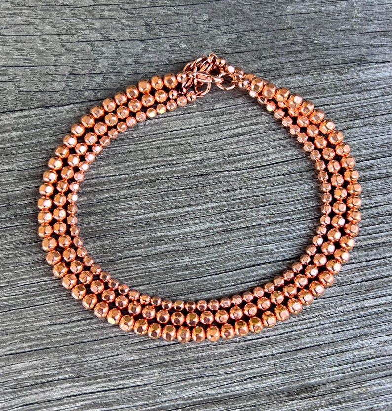 K&ouml;nnte beinhalten: Eine kupferfarbene Perlenkette mit dreireihigem Design. Die Halskette hat kleine, runde Perlen und einen passenden Verschluss. Der Schmuck wird auf einer Holzoberfl&auml;che pr&auml;sentiert.