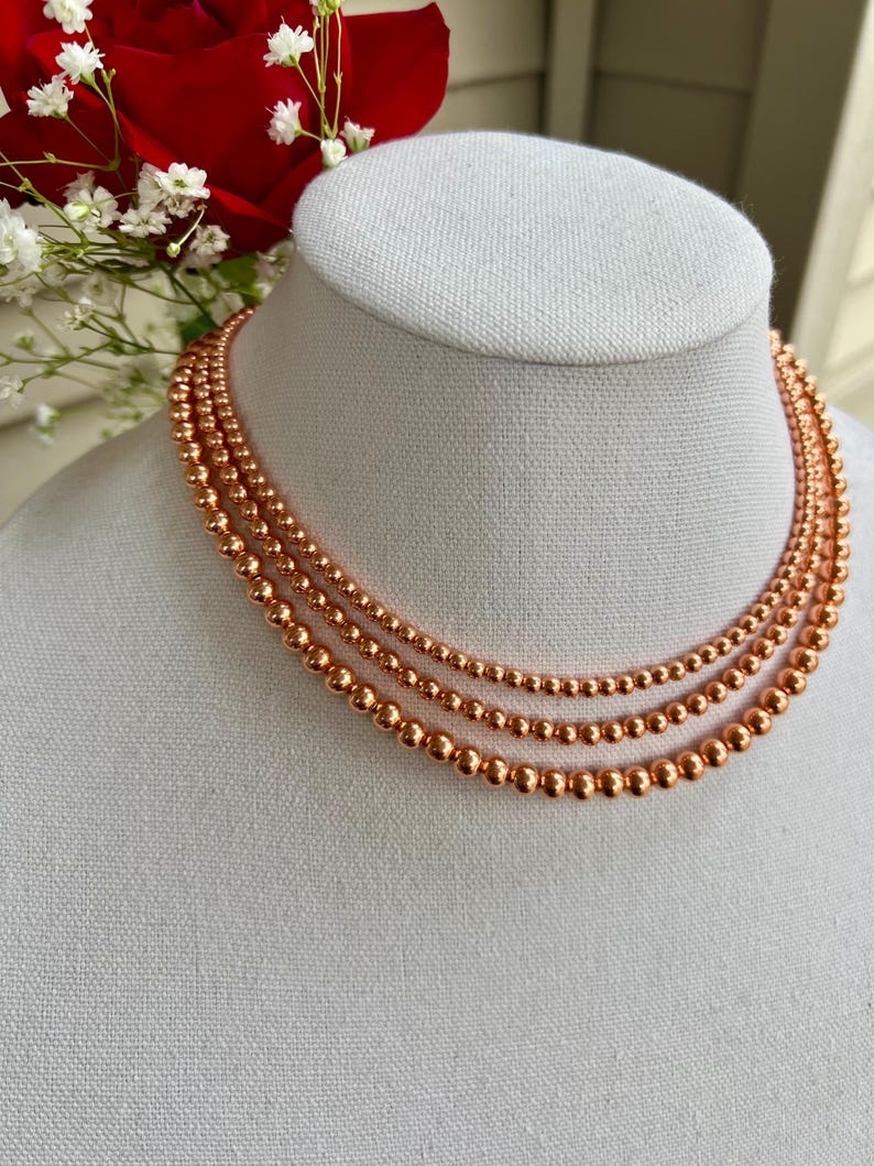 May include: A layered necklace made of three strands of copper beads. The beads are round and have a shiny, metallic finish.