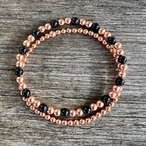 May include: Two beaded bracelets. One bracelet is made of small, round, rose gold beads. The second bracelet is made of rose gold beads and interspersed with black beads. The bracelets are displayed on a wooden surface.