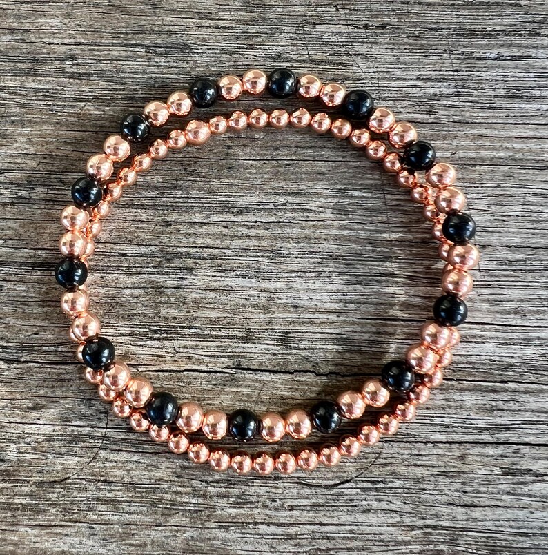 May include: Two beaded bracelets. One bracelet is made of small, round, rose gold beads. The second bracelet is made of rose gold beads and interspersed with black beads. The bracelets are displayed on a wooden surface.