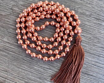 May include: A copper-colored mala bead necklace with a brown tassel. The necklace is made of small, round beads strung together. The tassel is made of multiple strands of brown thread. The necklace is displayed on a wooden surface.