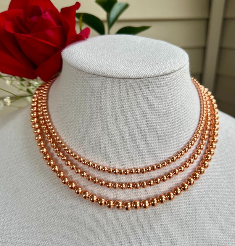 May include: A layered necklace made of three strands of copper beads. The beads are round and have a shiny, metallic finish.