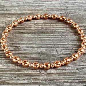May include: A copper-colored beaded bracelet. The bracelet is made of small, round beads strung together on an elastic band. The beads have a shiny, metallic finish. The bracelet is displayed on a wooden surface.