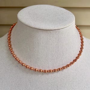 May include: A copper-coloured beaded necklace displayed on a white mannequin. The necklace is made up of small, evenly spaced beads, creating a delicate and elegant appearance. The beads have a metallic sheen, reflecting light.
