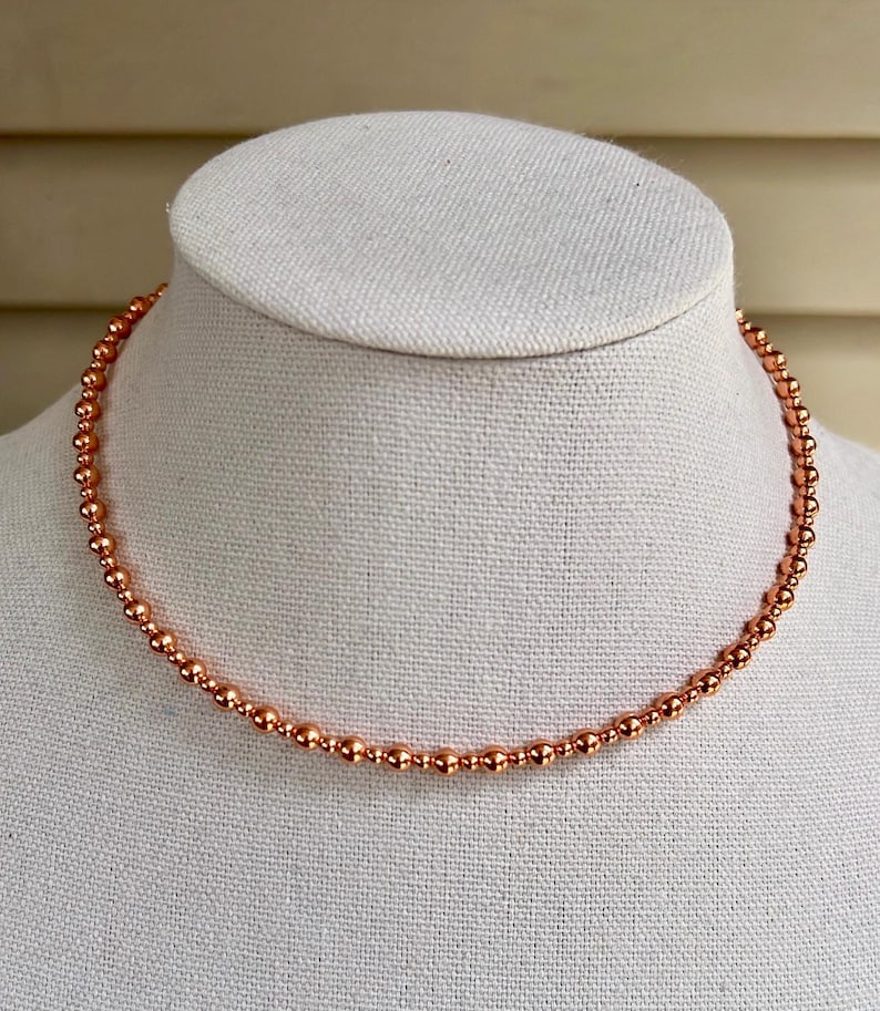May include: A copper-coloured beaded necklace displayed on a white mannequin. The necklace is made up of small, evenly spaced beads, creating a delicate and elegant appearance. The beads have a metallic sheen, reflecting light.