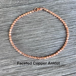 May include: A copper-colored faceted bead anklet. The anklet is made of small, round beads strung together, with a lobster clasp closure. The text "Faceted Copper Anklet" is printed below the anklet.