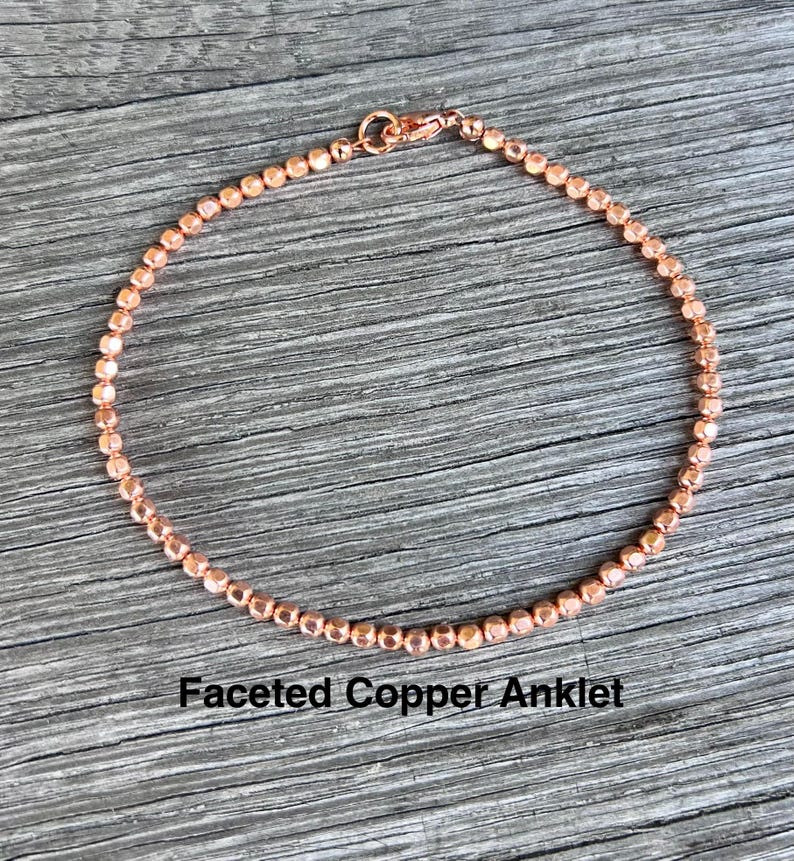K&ouml;nnte beinhalten: Ein kupferfarbenes Fu&szlig;kettchen mit facettierten Perlen. Das Fu&szlig;kettchen besteht aus kleinen, runden Perlen, die aneinandergereiht sind, mit einem Karabinerverschluss. Der Text "Faceted Copper Anklet" ist darunter gedruckt.
