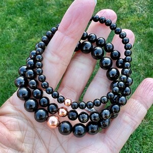 May include: Three black beaded bracelets with rose gold accents. The bracelets are layered on top of each other and are held in a hand.
