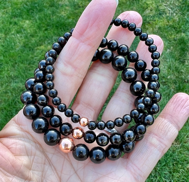 May include: Three black beaded bracelets with rose gold accents. The bracelets are layered on top of each other and are held in a hand.