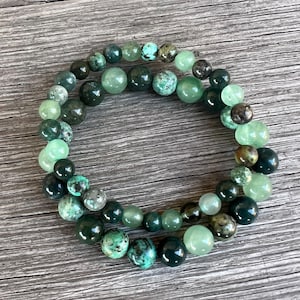 May include: Two beaded bracelets with a variety of green gemstones. The beads range in color from light mint to deep forest green, with some featuring turquoise-colored speckles. The bracelets are displayed on a wooden surface.