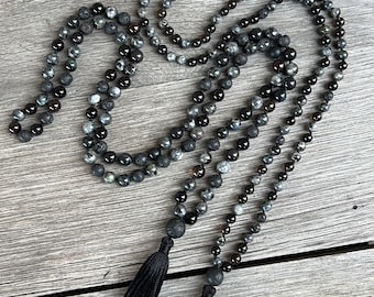 May include: Two beaded necklaces with black tassels, featuring dark grey, black, and speckled grey beads. The necklaces are displayed on a weathered, grey wooden surface.