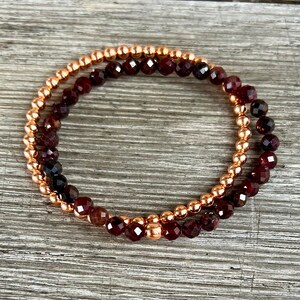 May include: Two stacked bracelets. The top bracelet features small, round, rose gold beads. The bottom bracelet has faceted, dark red beads. Both bracelets are on a light brown wooden surface.