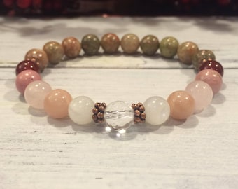 May include: A bracelet made with various semi-precious stones, including pink, red, and green, with a clear quartz crystal bead in the center. The bracelet is accented with copper-colored beads.
