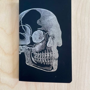 May include: A black notebook featuring a detailed, white illustration of a human skull in profile. The anatomical drawing is a stark contrast against the dark background, highlighting the skull's structure.