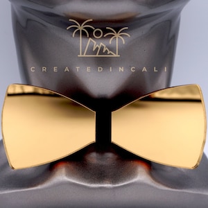 Puede incluir: Una pajarita dorada con superficie reflectante. La pajarita se muestra en una cabeza de maniquí gris con el texto "CREATED IN CALI" impreso en la parte delantera.