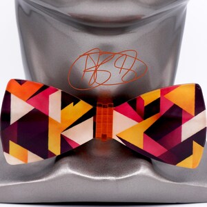 May include: A bow tie featuring a geometric pattern of triangles in shades of orange, yellow, magenta, and deep purple. The center knot is a translucent orange plastic. The bow tie is displayed on a metallic gray mannequin head with a signature.