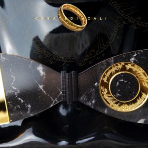 May include: A black and gold bow tie with a marble pattern and a gold ring with the inscription "One Ring to rule them all" on the front. The inscription is in Elvish.