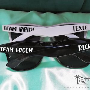 May include: Two pairs of sunglasses with black frames and dark lenses. One pair has white frames with the text "TEAM BRIDE" and "LEXIE" printed on them. The other pair has black frames with the text "TEAM GROOM" and "RICK" printed on them.