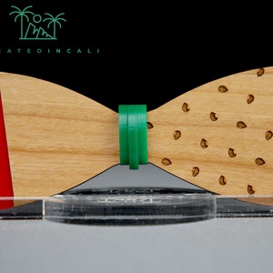 Pode incluir: Uma gravata borboleta de madeira com um padrão de madeira vermelha e marrom em um lado e um padrão de madeira marrom com pequenos pontos marrons no outro lado. A gravata borboleta é mantida unida por uma banda elástica verde.