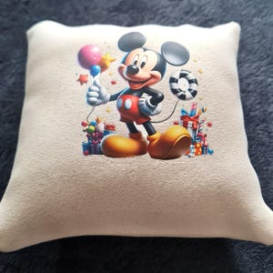 Può includere: Un cuscino quadrato bianco sporco con un'immagine colorata di Topolino che tiene in mano un palloncino e una stella. Mickey indossa pantaloncini rossi, scarpe gialle e guanti bianchi. Anche regali e caramelle sono nell'immagine.