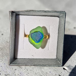 Puede incluir: Un anillo de tono dorado con un diseño en capas que presenta una pieza de vidrio marino verde lima translúcido rematada por una pieza más pequeña de vidrio marino azul profundo. El anillo se presenta dentro de una caja de joyería cuadrada gris.